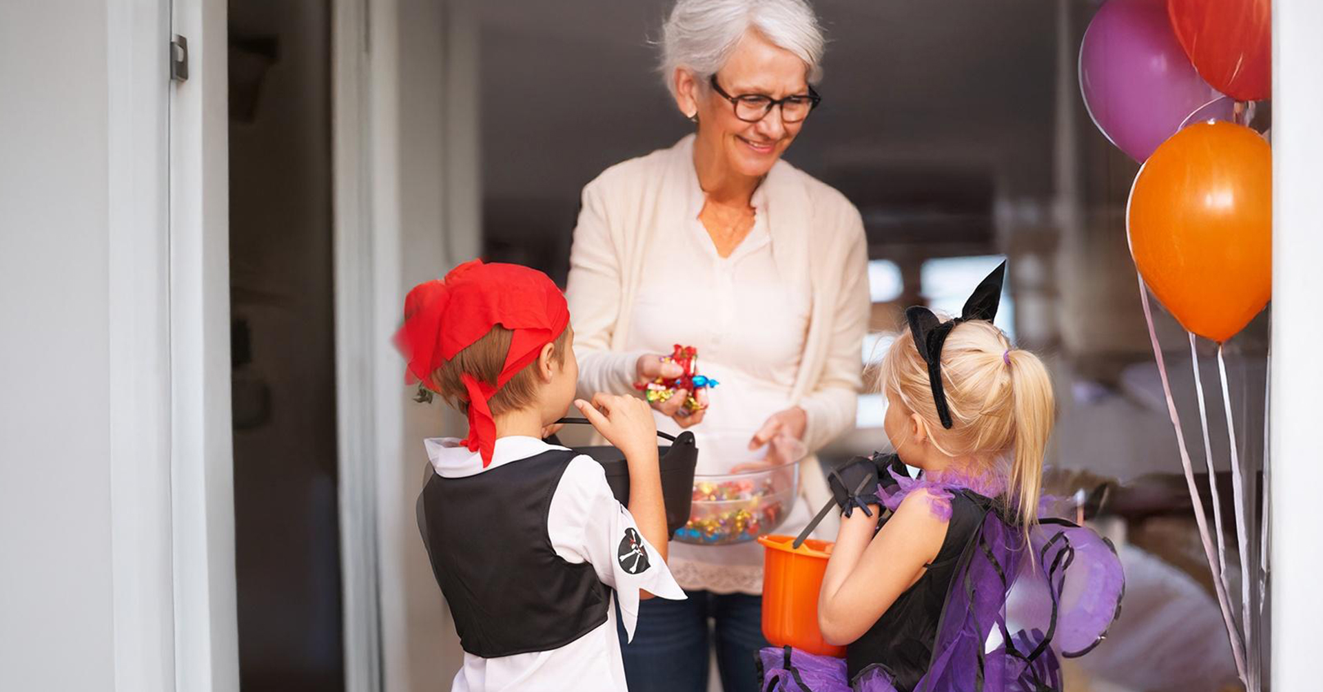 senior woman handing out candy to 2 kids trick-or-treating -- one dressed up as a pirate, and the other dressed up as a bat