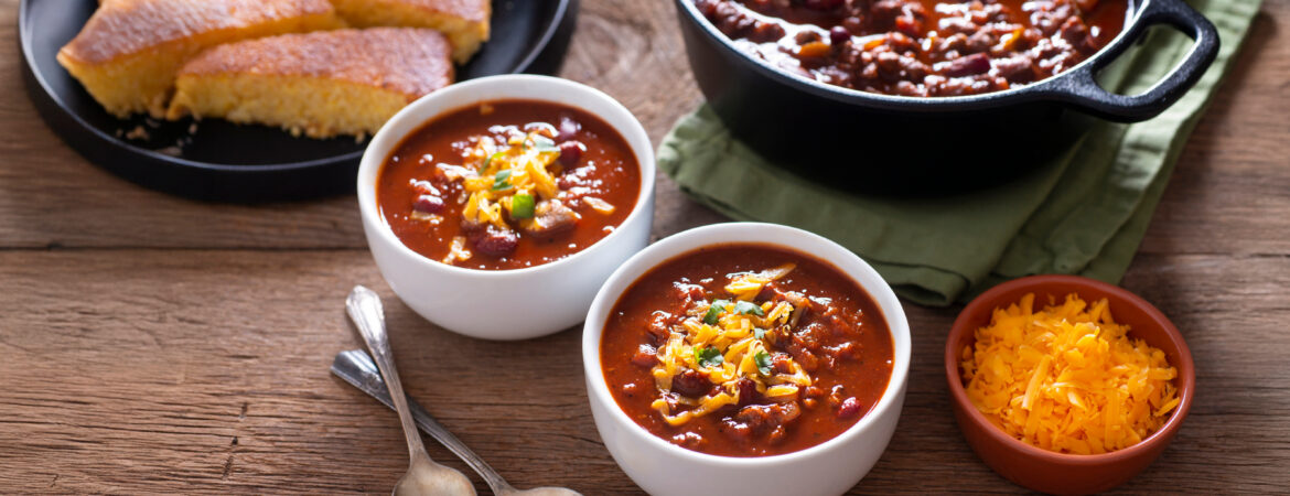 A pot of chili next to a plate of cornbread, and 2 bowls of chili with shredded cheese on top.