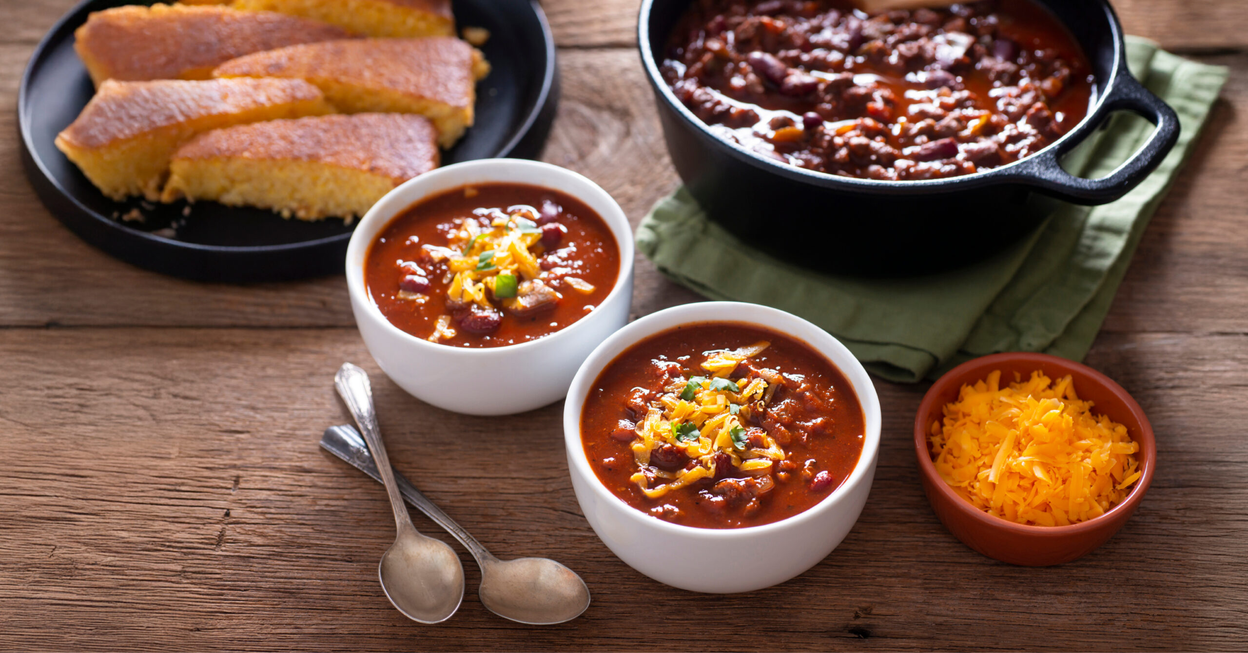 A pot of chili next to a plate of cornbread, and 2 bowls of chili with shredded cheese on top.