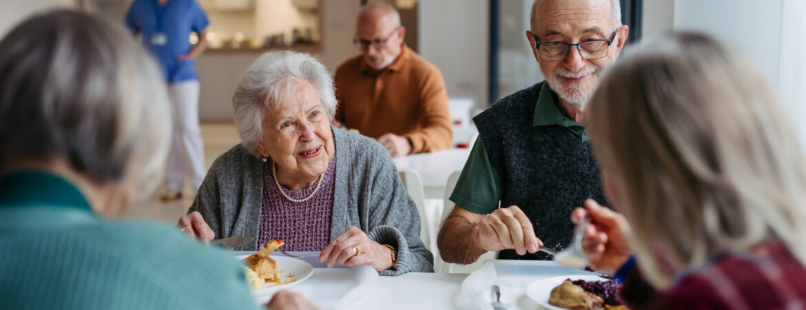 Older adults are dining together at a bright, communal table, smiling and engaged in conversation, creating a warm and friendly atmosphere eating pie.