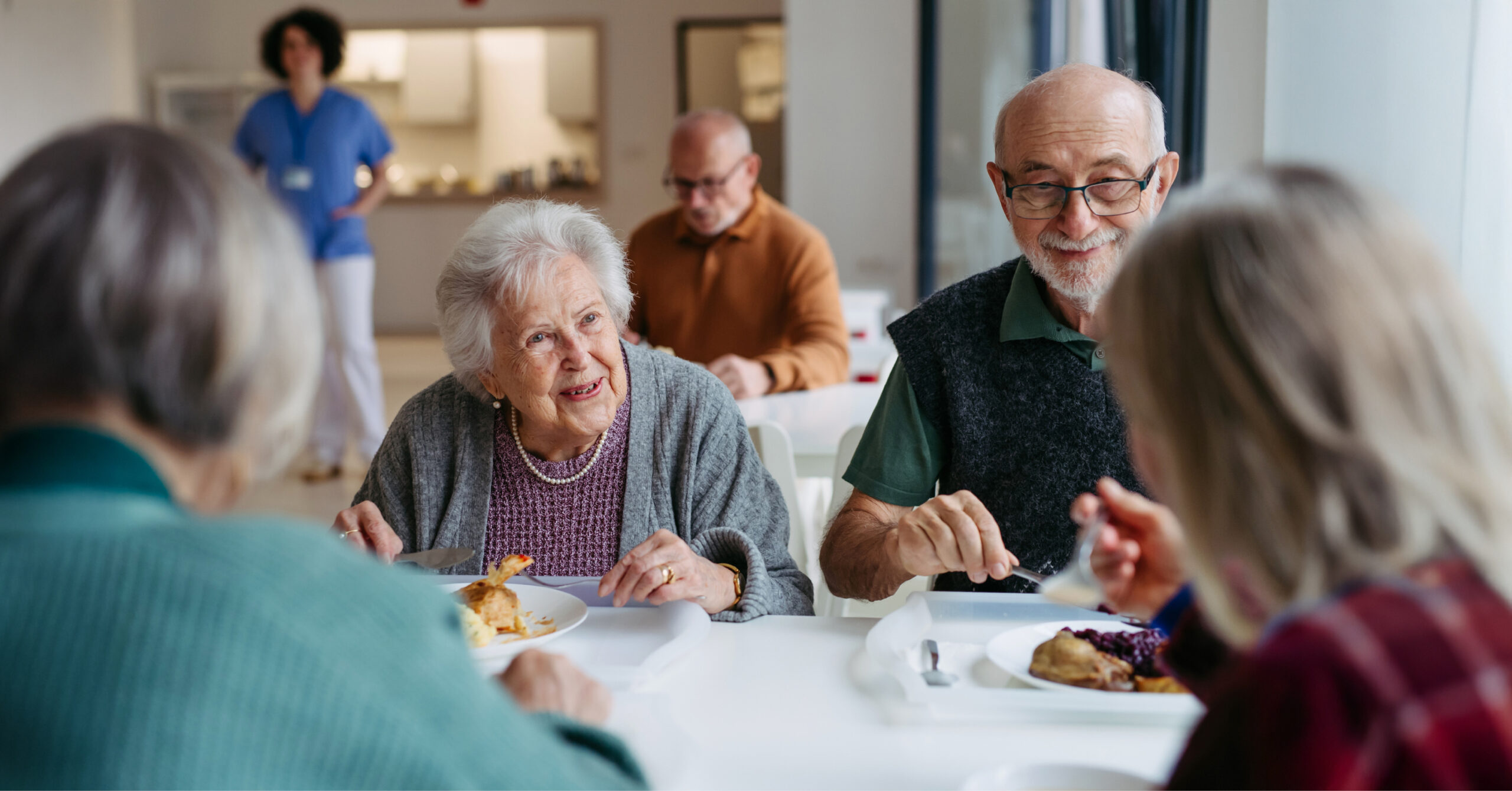 Older adults are dining together at a bright, communal table, smiling and engaged in conversation, creating a warm and friendly atmosphere eating pie.