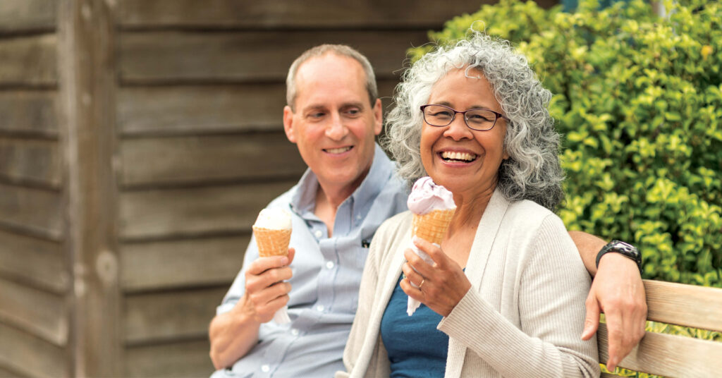 A smiling elderly couple sits on a bench, enjoying ice cream cones. The woman laughs joyfully, and the man looks at her warmly. Background features wooden siding and greenery.